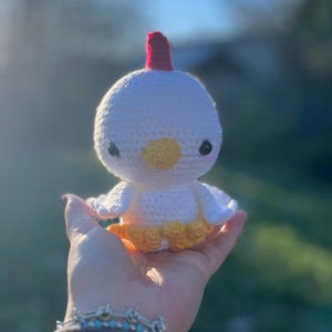 May include: A handmade white crochet chicken with a red comb, yellow beak, and orange skirt. The amigurumi chicken has black eyes and is held in a hand against a blurred background. The crochet chicken is a cute, whimsical toy.