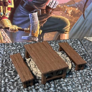 May include: Miniature wooden table and benches set. The table has a rectangular top and four legs. Two benches are also included. The set is made of brown wood and is designed for tabletop gaming or miniature displays.