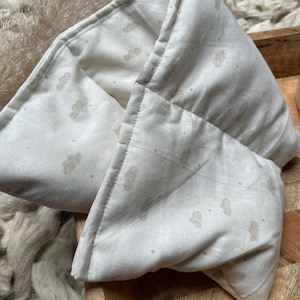 Peut inclure: Un enveloppement de cou rectangulaire de couleur crème avec un motif de nuages. L'enveloppement est plié et semble être fait de tissu doux. Le motif de nuages est de couleur marron clair. L'enveloppement repose sur un plateau en bois.