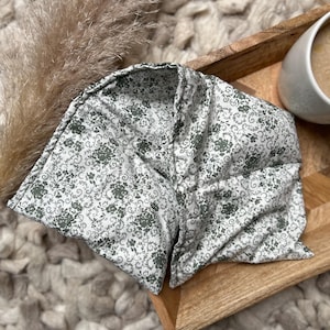 Peut inclure: Un tour de cou à motifs floraux blancs et verts, posé sur un plateau en bois. Le tissu présente un motif délicat. Une tasse de café est visible en arrière-plan.
