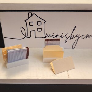 May include: Miniature office supplies set, including file folders and lined paper, displayed on a white surface. The background features a white sign with a house outline and the text "minisbycm" in black script.