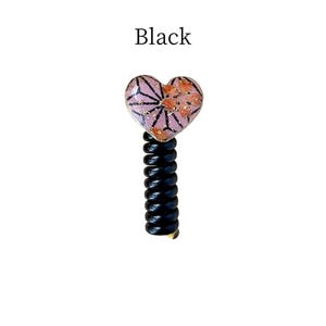May include: A black hair accessory with a heart-shaped top. The heart has a pink and orange floral design with black outlines. The text "Black" is above the accessory.