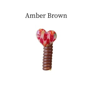 May include: A heart-shaped decorative object with a spring base. The heart is red with gold and white accents. The spring base is brown, and the text "Amber Brown" is above the object.