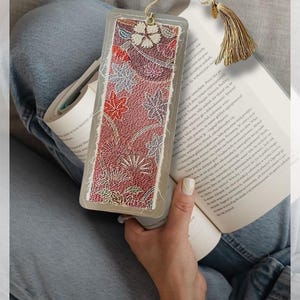 以下が含まれることがあります： 赤、白、青の色合いの花柄が特徴の長方形のブックマーク。ブックマークには金のタッセルと透明なプラスチックケースが付いています。本の中にあります。