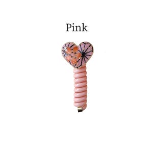 May include: A pink heart-shaped phone cord with a floral design and the word "Pink". The heart has a pink background with orange and black accents. The cord is a light pink colour.
