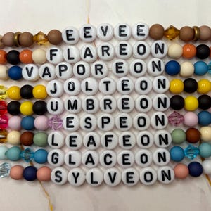 Könnte beinhalten: Perlenaccessoires mit weißen Buchstabenperlen, die "EEVEE", "FLAREON", "VAPOREON", "JOLTEON", "UMBREON", "ESPEON", "LEAFEON", "GLACEON" und "SYLVEON" buchstabieren. Die Perlen sind in verschiedenen Farben, darunter Braun, Orange, Blau und Rosa.