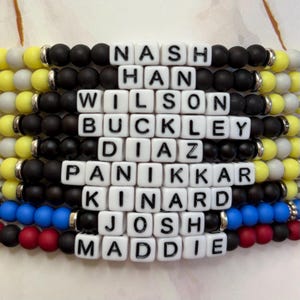 Könnte beinhalten: Ein Perlenarmband mit weißen Buchstabenperlen, die Namen wie Nash, Han und Wilson buchstabieren. Das Armband hat schwarze, gelbe, rote und blaue Perlen mit silbernen Akzenten. Die Namen sind auf dem Armband zentriert.