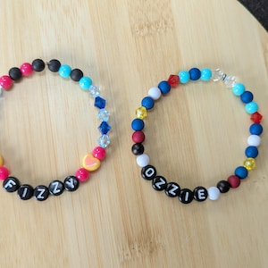 Könnte beinhalten: Zwei Armbänder mit bunten Perlen und Buchstabenperlen mit den Wörtern "FIZZY" und "OZZIE". Die Armbänder haben eine Vielzahl von Farben, darunter Rot, Blau, Schwarz, Weiß und Gelb, und werden auf einer Holzoberfläche präsentiert.