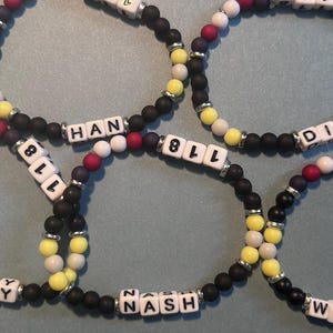 Könnte beinhalten: Mehrere Armbänder mit Perlen, auf denen weiße Buchstabenperlen Namen und Zahlen bilden. Die Armbänder haben schwarze, gelbe, weiße und rote Perlen, die durch silberfarbene Abstandshalter getrennt sind. Die Namen "BUCKLEY", "HAN", "DIAZ", "NASH" und "WILSON" sind sichtbar.