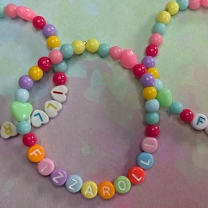 Könnte beinhalten: Drei farbenfrohe Armbänder mit Perlen, auf denen der Name "FIZZAROL" mit weißen Buchstabenperlen geschrieben steht. Die Armbänder haben eine Vielzahl von pastellfarbenen Perlen, darunter rosa, gelb, blau und grün, mit herzförmigen Perlen.