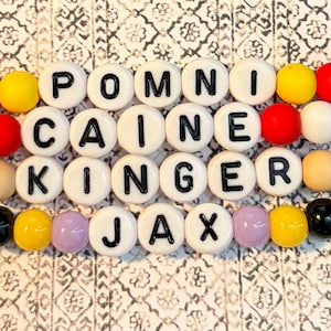 Könnte beinhalten: Ein Armband mit weißen Buchstabenperlen, die die Namen POMNI, CAINE, KINGER und JAX buchstabieren. Die Perlen sind mit bunten runden Perlen in Rot, Gelb, Blau, Grün und anderen Farben durchsetzt. Der Hintergrund ist ein gemusterter Stoff.