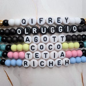 Könnte beinhalten: Sechs gestapelte Perlenarmbänder mit runden weißen Perlen und schwarzen Buchstaben, die QIFREY, OLRUGGIO, AGOTT, COCO, TETIA und RICHEH bilden. Die Armbänder enthalten verschiedenfarbige Perlen in Schwarz, Weiß, Gelb, Rosa und Hellblau sowie goldene Akzente.