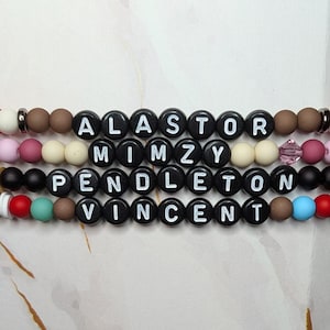 Könnte beinhalten: Nahaufnahme von vier Armbändern mit Perlen. Jedes Armband hat schwarze runde Perlen mit weißen Buchstaben, die die Namen Alastor, Mimzy, Pendleton und Vincent buchstabieren. Die Armbänder haben verschiedenfarbige Perlen und Akzente.