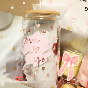 May include: A Valentine's Day gift box filled with a glass tumbler with a bamboo lid and straw, a candle with a pink bow sticker, a chocolate candy, and tissue paper with gold and pink hearts. The tumbler has a pink and gold sticker that says "love you".