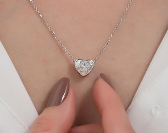 Puede incluir: Un delicado collar de plata con un colgante de diamante en forma de corazón. El colgante es una piedra preciosa clara y brillante, suspendida de una fina cadena. El collar se muestra sobre un fondo suave y neutro, resaltando la elegancia de la joya.