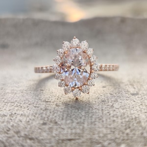 May include: Rose gold engagement ring featuring an oval-cut center stone surrounded by a halo of smaller diamonds. The band is also adorned with small diamonds, creating a sparkling effect. The ring is set against a neutral background, highlighting its elegant design.
