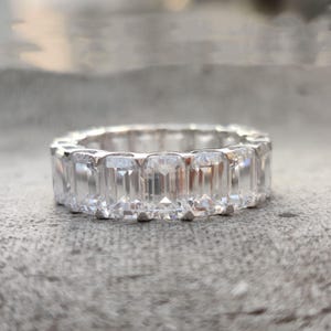 May include: A silver eternity ring featuring a continuous row of emerald-cut, clear gemstones. The ring is set on a gray surface, showcasing the sparkle of the stones. This piece is a classic jewelry item.