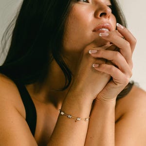 Puede incluir: Un brazalete de oro con una cadena delicada y pequeñas piedras cuadradas transparentes.