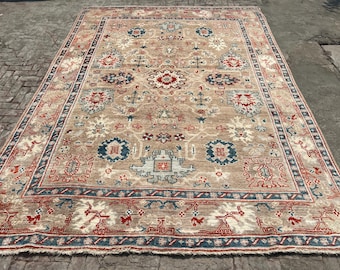 Könnte beinhalten: Ein großer, rechteckiger Teppich mit einem aufwendigen, traditionellen Design. Der Teppich hat einen beigefarbenen Hintergrund mit einem sich wiederholenden Muster aus roten, blauen und cremefarbenen floralen und geometrischen Motiven. Ein blauer Rand umrahmt den Teppich.