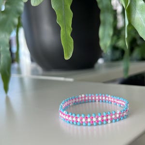 Puede incluir: Un brazalete de cuentas azul, blanco y rosa. El brazalete está hecho de pequeñas cuentas redondas que están enhebradas juntas en un patrón.