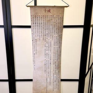 May include: A rectangular scroll with Japanese text, hanging from a wooden rod. The text is written in vertical columns and appears to be a religious or philosophical text. The scroll is beige with black text.