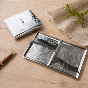 May include: Two silver-toned metal cigarette cases with an intricate embossed floral pattern. One case is closed, displaying the name Polli in script, while the other is open, revealing two internal metal spring-loaded bars for securing contents.