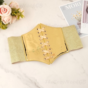 May include: A gold-colored corset belt with a shiny, metallic finish. The belt features a lace-up design in the center with satin ribbon and gold elastic side panels. The belt is a fashion accessory.