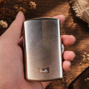 May include: A rectangular silver-toned metal cigarette case featuring a fine herringbone texture across the surface. The bottom center of the case displays the word Polli engraved within a rectangular frame. The case has a hinged side closure.