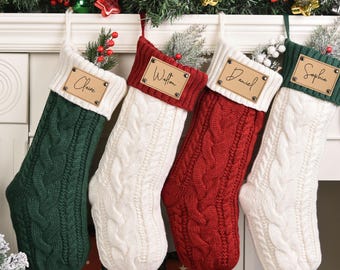 以下が含まれることがあります： 緑、白、赤のケーブルニットのクリスマスストッキングが4つ、白いマントルピースから吊り下げられています。各ストッキングには、パーソナライズされた木製のネームタグが付いています。お祝いのガーランドとクリスマスツリーが背景にあります。