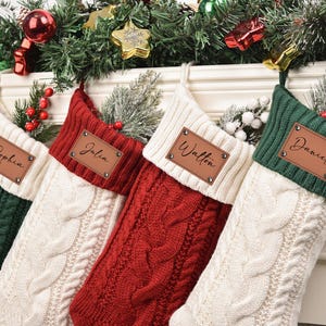 May include: Four personalized Christmas stockings in green, red, and cream colors, each with a leather name tag. The stockings are cable-knit and decorated with festive greenery and ornaments, perfect for holiday decor.