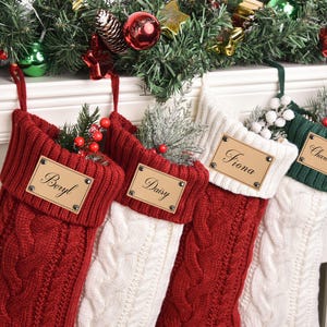 Op de afbeelding: Vier kabelgebreide kerstsokken in rood, wit en groen, elk met een gepersonaliseerd naamlabel. De sokken zijn versierd met groen en bessen en hangen aan een schoorsteenmantel met kerstversieringen.