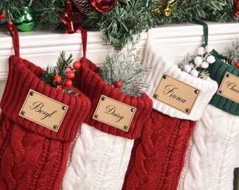 以下が含まれることがあります： 赤、白、緑のケーブルニットのクリスマスストッキング4つ。それぞれにパーソナライズされたネームタグが付いています。ストッキングは緑とベリーで飾られ、クリスマスの飾りがあるマントルピースから吊り下げられています。