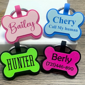 May include: Four colorful bone-shaped dog tags with personalized names and phone numbers. The tags are pink, blue, green, and hot pink. The names on the tags are Bailey, Chery, Hunter, and Berly. The phone number on the hot pink tag is (721) 446-890. The blue tag also has the text "Call My Human".