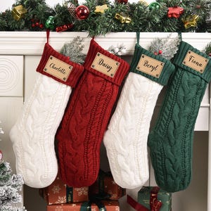 Op de afbeelding: Vier kabelgebreide kerstsokken in wit, rood en groen hangen boven een open haard. Elke sok heeft een gepersonaliseerd naamlabel: Charlotte, Daisy, Beryl en Fiona. Feestelijk groen en ornamenten sieren de schoorsteenmantel.
