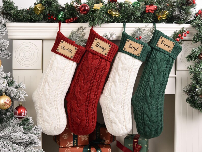 Op de afbeelding: Vier kabelgebreide kerstsokken in wit, rood en groen hangen boven een open haard. Elke sok heeft een gepersonaliseerd naamlabel: Charlotte, Daisy, Beryl en Fiona. Feestelijk groen en ornamenten sieren de schoorsteenmantel.