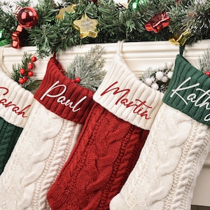 May include: Four cable-knit Christmas stockings in red, white, and green, each with a name embroidered in a contrasting colour. The stockings are decorated with festive greenery and berries, ideal for holiday decor.