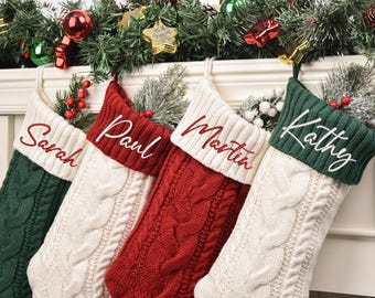 Op de afbeelding: Vier kabelgebreide kerstsokken in rood, wit en groen, elk met een naam geborduurd in een contrasterende kleur. De sokken zijn versierd met feestelijk groen en bessen, perfect voor vakantiedoeleinden.