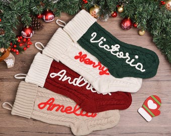 May include: Four cable-knit Christmas stockings with ribbed cuffs and hanging loops. The top stocking is dark green with white text reading Victoria. The second is cream with red text reading Henry. The third is red with white text reading Andrew. The bottom is beige with red text reading Amelia.