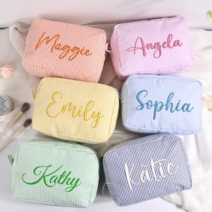 May include: Six colourful, striped cosmetic bags with embroidered names: Maggie, Angela, Emily, Sophia, Kathy, and Katie. The bags are in shades of orange, pink, yellow, blue, green, and navy, with white stripes. Each bag has a zip closure.