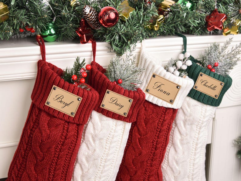 Op de afbeelding: Vier kabelgebreide kerstsokken in rood, wit en groen, elk met een gepersonaliseerd naamlabel. De sokken zijn versierd met groen en bessen en hangen aan een schoorsteenmantel met kerstversieringen.