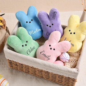 May include: A wicker basket holds six plush bunny-shaped toys in pastel colors: blue, green, purple, pink, and yellow. Each bunny has black eyes and a name or year embroidered on it. Easter-themed decor.
