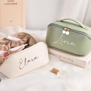 May include: Two cosmetic bags, one open and one closed. The open bag is cream-coloured and contains makeup products. The closed bag is sage green with a handle and the name "Liora" in script. Both bags have gold zips.