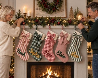 Op de afbeelding: Vijf hangende kerstsokken met gepersonaliseerde namen in cursief schrift. De sokken zijn uitgevoerd in rood-witte strepen, groen-witte ruitjes, rood-witte ruitjes, effen oudroze en groen-witte strepen, elk voorzien van een decoratieve strik.