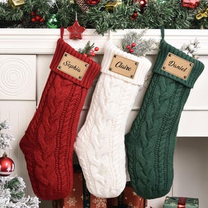 Op de afbeelding: Drie kabelgebreide kerstsokken in rood, wit en groen hangen aan een witte schoorsteenmantel. Elke sok heeft een leren label met een naam: Sophia, Claire en Daniel. Feestelijke vakantiedecoratie.
