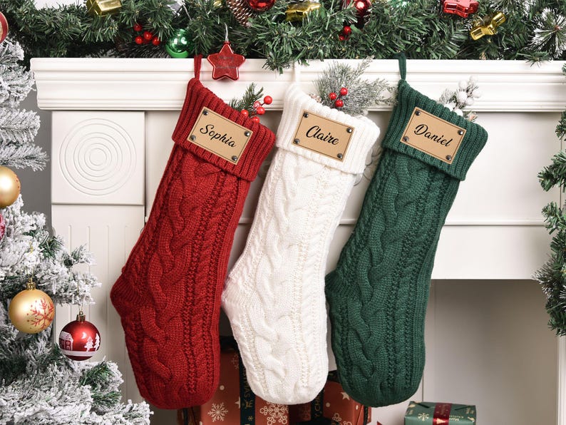 Op de afbeelding: Drie kabelgebreide kerstsokken in rood, wit en groen hangen aan een witte schoorsteenmantel. Elke sok heeft een leren label met een naam: Sophia, Claire en Daniel. Feestelijke vakantiedecoratie.