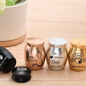 Puede incluir: Una colección de pequeñas urnas personalizadas en varios acabados metálicos. Una urna negra tiene el nombre "Baldie" y huellas de patas. Otras urnas están grabadas con nombres, fechas y frases como "Love You Forever" y "God Holds You".