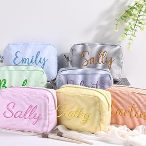 Peut inclure: Plusieurs trousses de maquillage personnalisées de différentes couleurs, dont bleu, vert, rose, jaune et orange. Chaque trousse présente un motif à rayures verticales et un nom brodé dans une couleur complémentaire. Les trousses sont zippées et semblent être en tissu texturé.