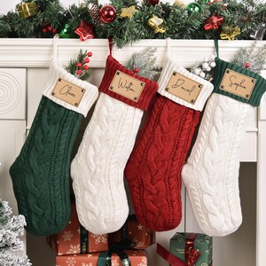 Op de afbeelding: Vier kabelgebreide kerstsokken in groen, wit en rood hangen aan een witte schoorsteenmantel. Elke sok heeft een gepersonaliseerd houten naamlabel. Feestelijke guirlande en kerstboom op de achtergrond.