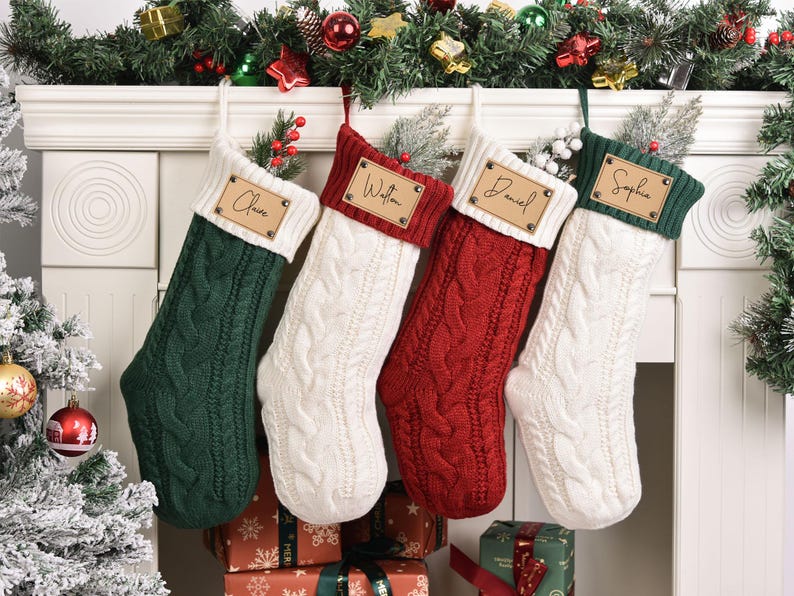 Op de afbeelding: Vier kabelgebreide kerstsokken in groen, wit en rood hangen aan een witte schoorsteenmantel. Elke sok heeft een gepersonaliseerd houten naamlabel. Feestelijke guirlande en kerstboom op de achtergrond.