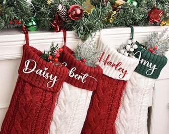 Op de afbeelding: Vier gepersonaliseerde kerstsokken in rood, wit en groen, met namen geborduurd in wit script. De sokken zijn kabelgebreid en versierd met groen en bessen, perfect voor de feestdagen.